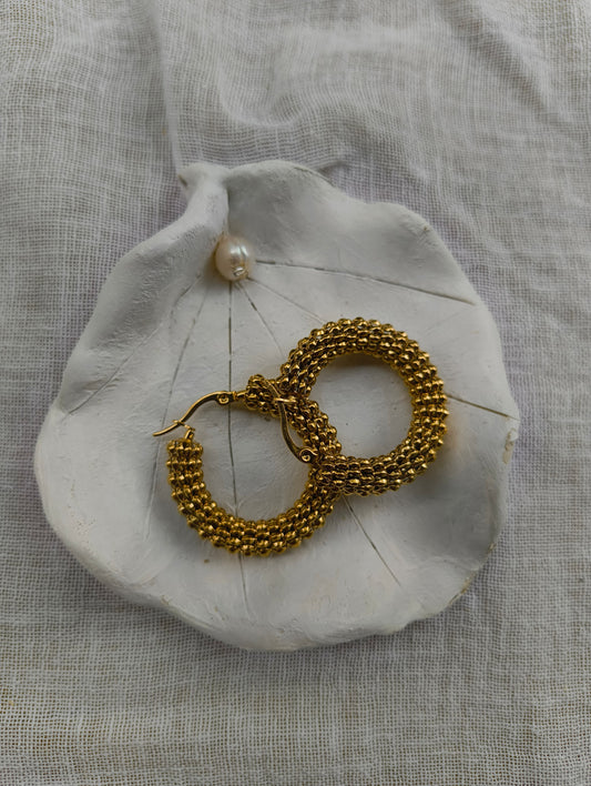 Gold hoop earrings on a white shell with a pearl, against a textured beige fabric background.