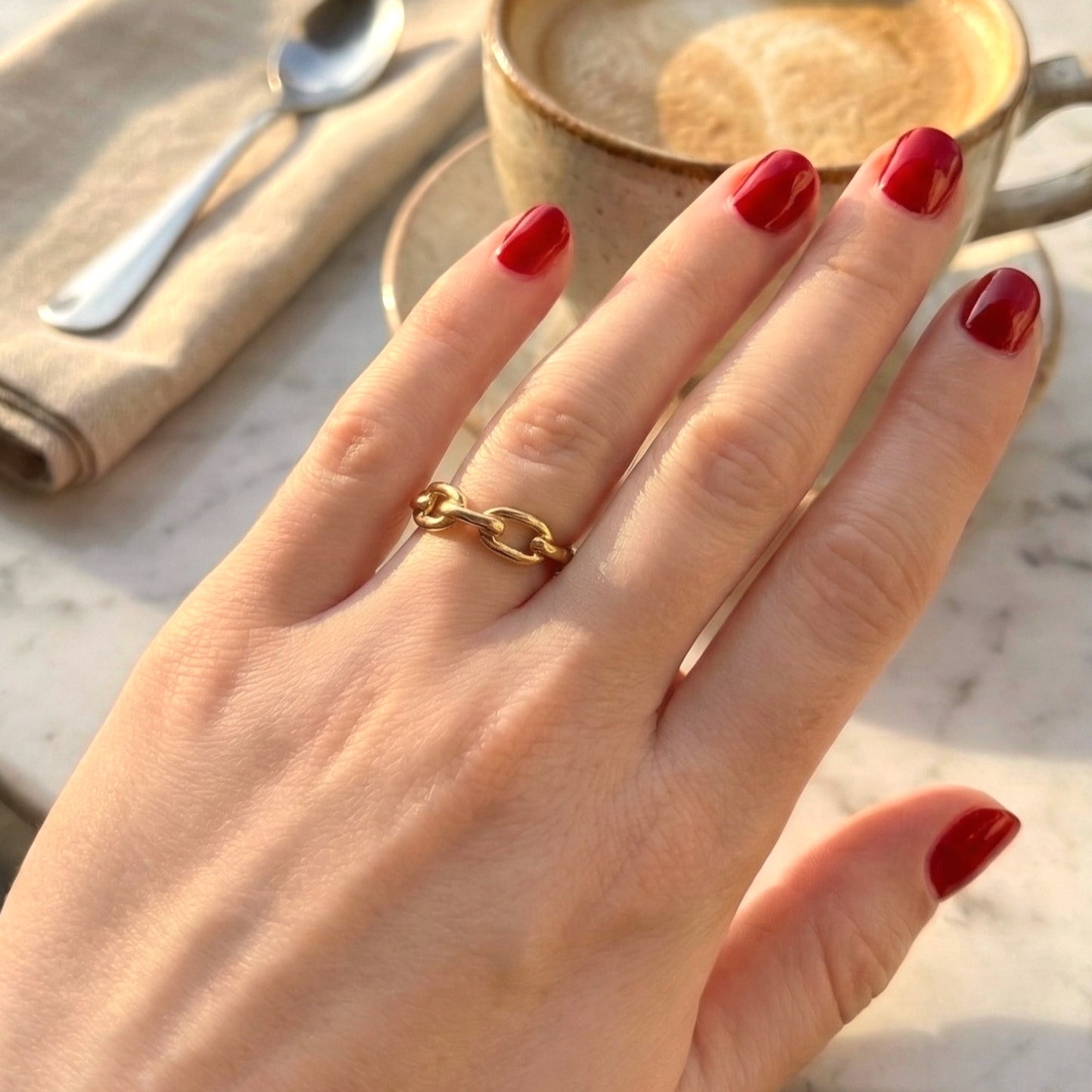 Hand wearing a gold ring with red nail polish, set against a blurred background of a cup and spoon.