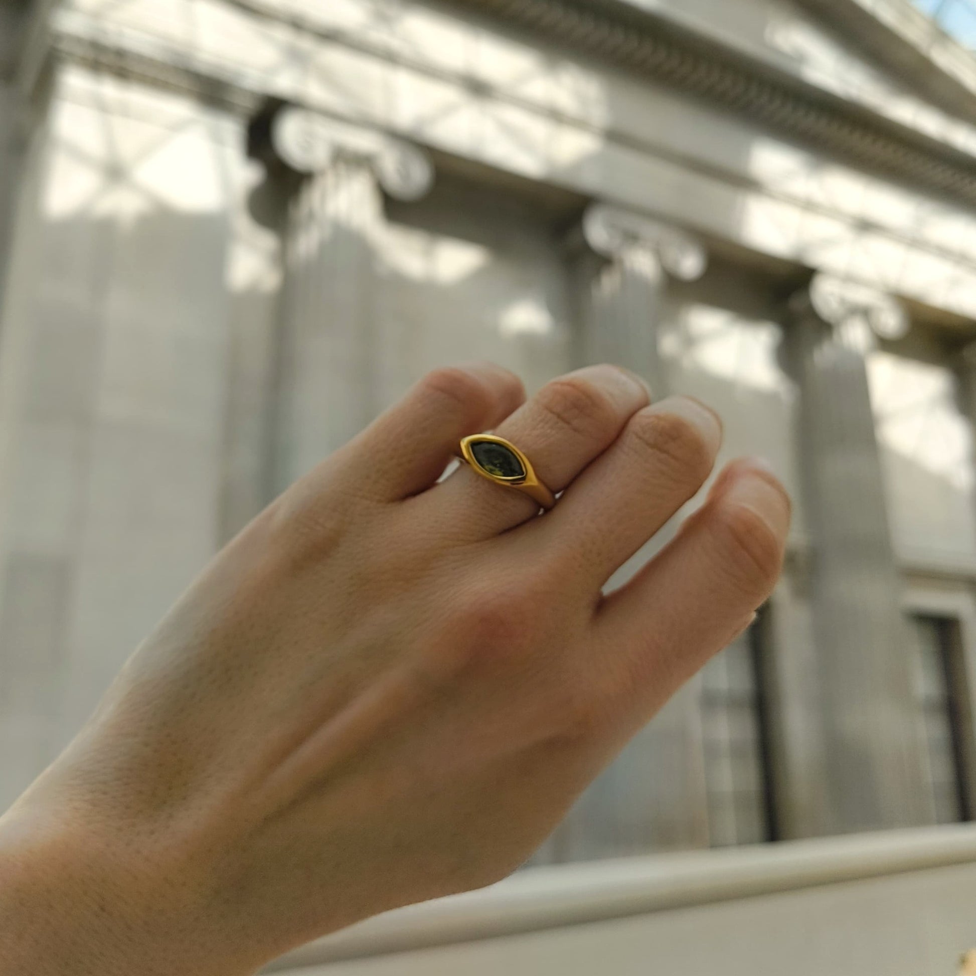 Hand wearing a gold ring with a green gemstone in front of a classical building.