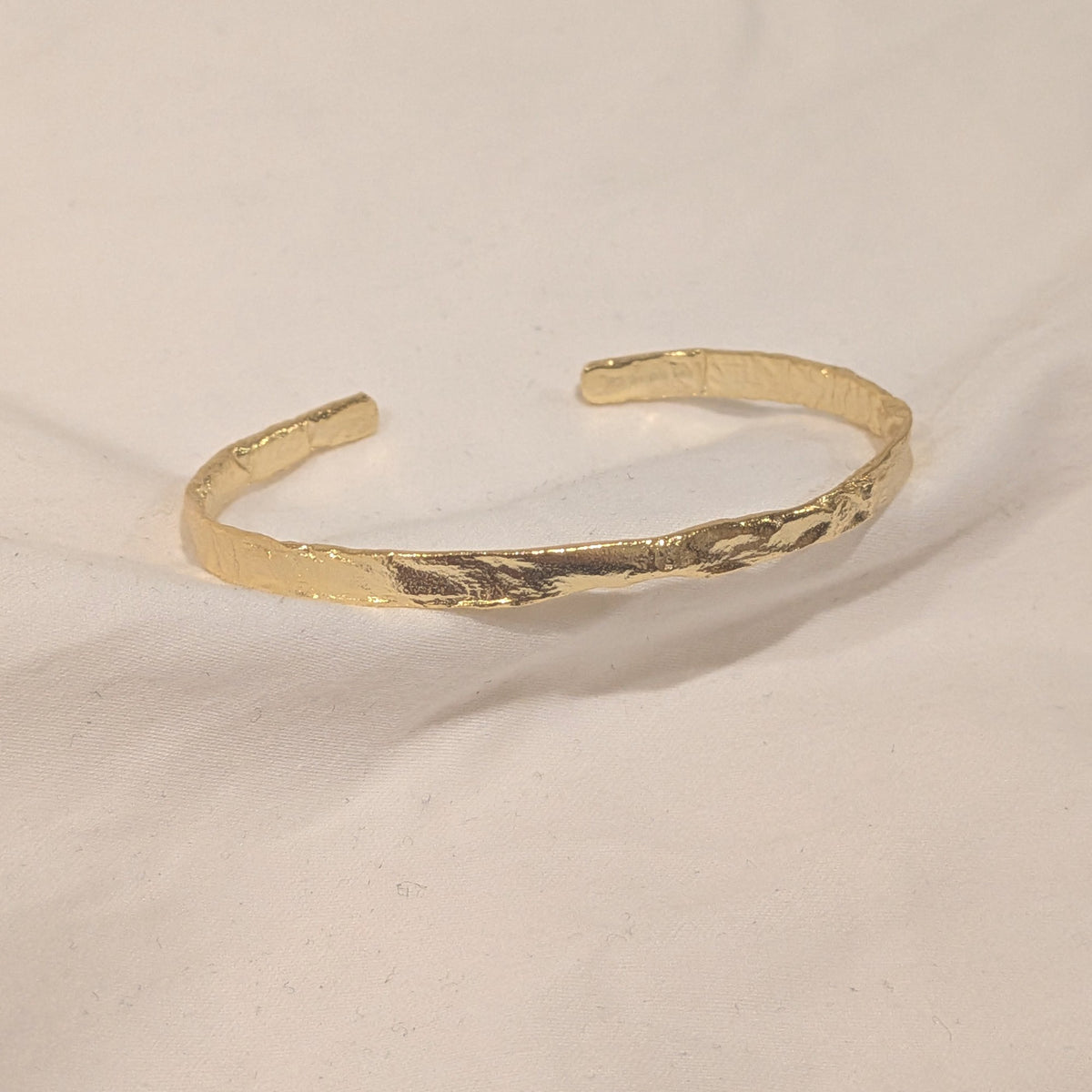 A textured gold open cuff bracelet with a hammered finish, displayed on a light beige fabric background.