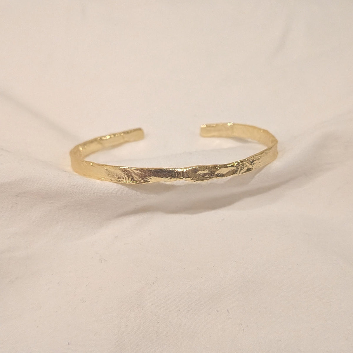 A textured gold open cuff bracelet resting on a white fabric background.