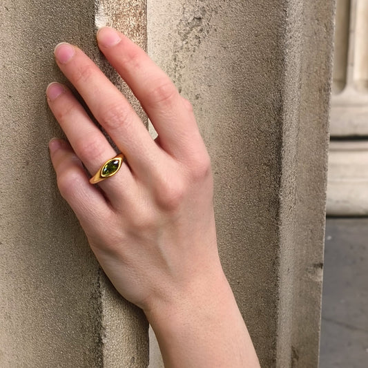 Hand wearing a gold ring with a green gemstone against a textured wall.