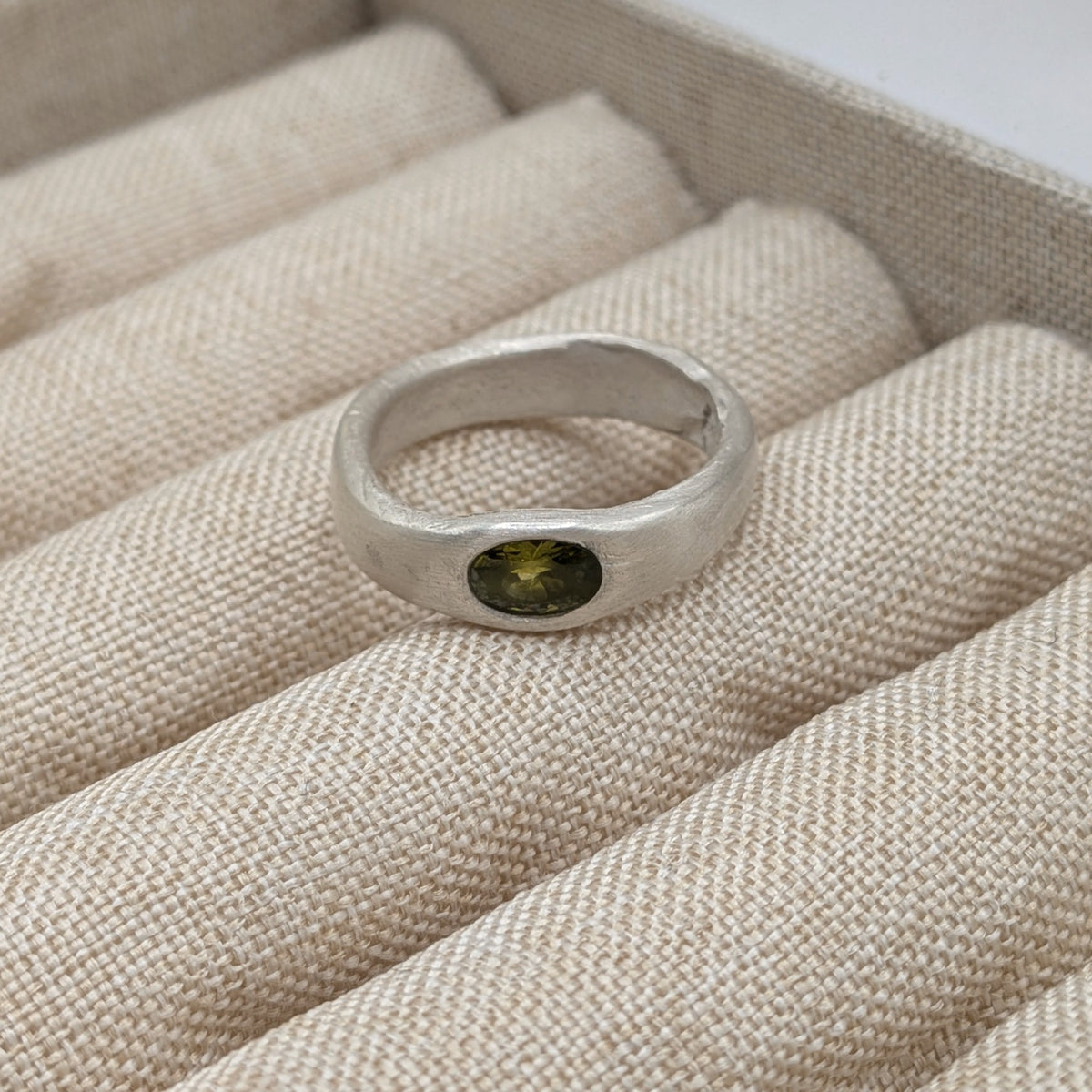A silver ring with a small oval green gemstone, displayed on a beige fabric ring box.