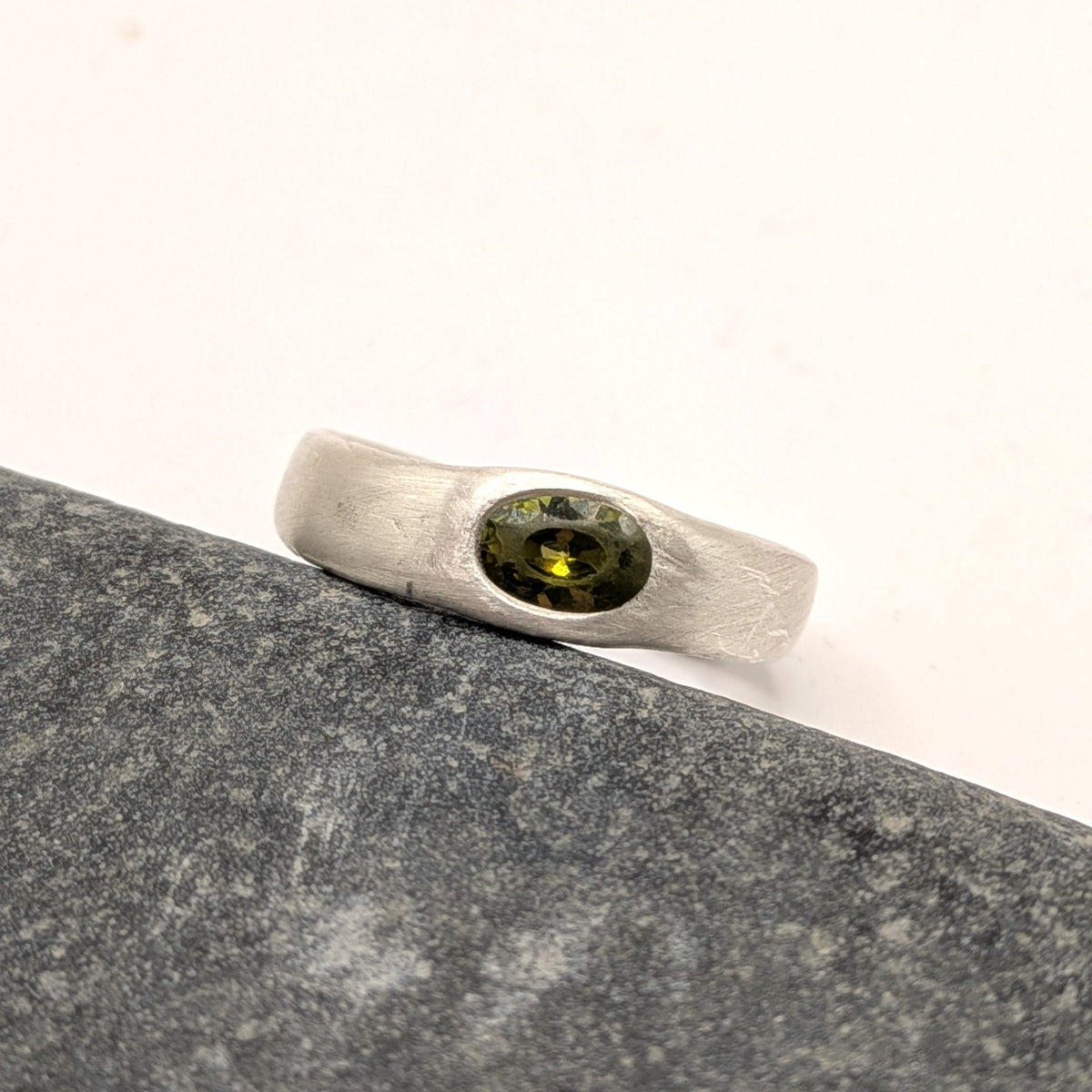 A textured sterling silver ring with a central oval green gemstone, resting on a dark gray stone slab against a white background.