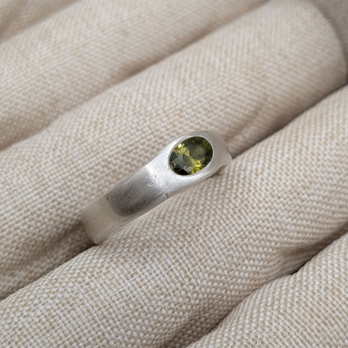 A sterling silver ring with a bezel-set oval green gemstone, displayed on a textured beige fabric ring holder.