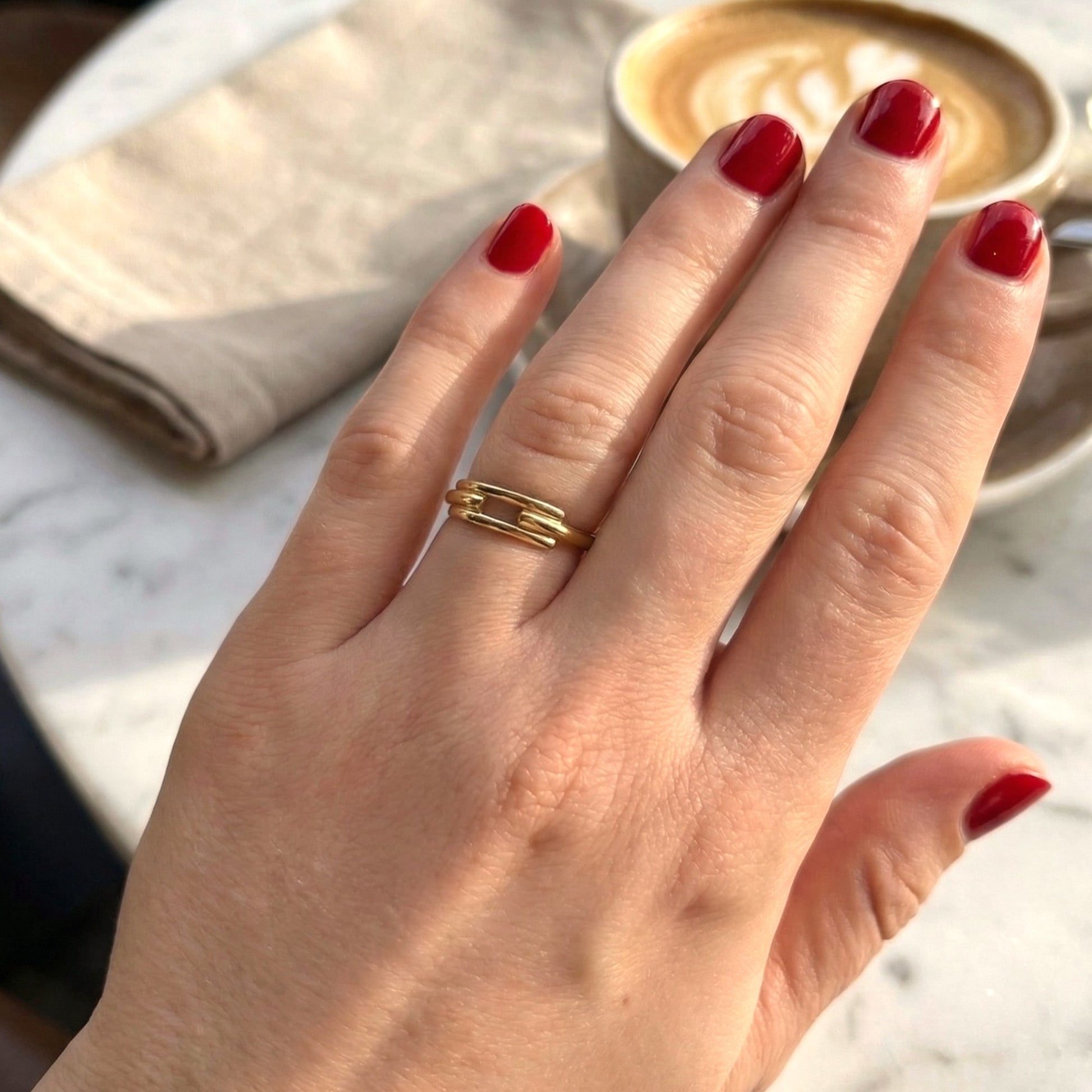 Hand with red nail polish wearing a gold ring, blurred background with a cup of coffee.