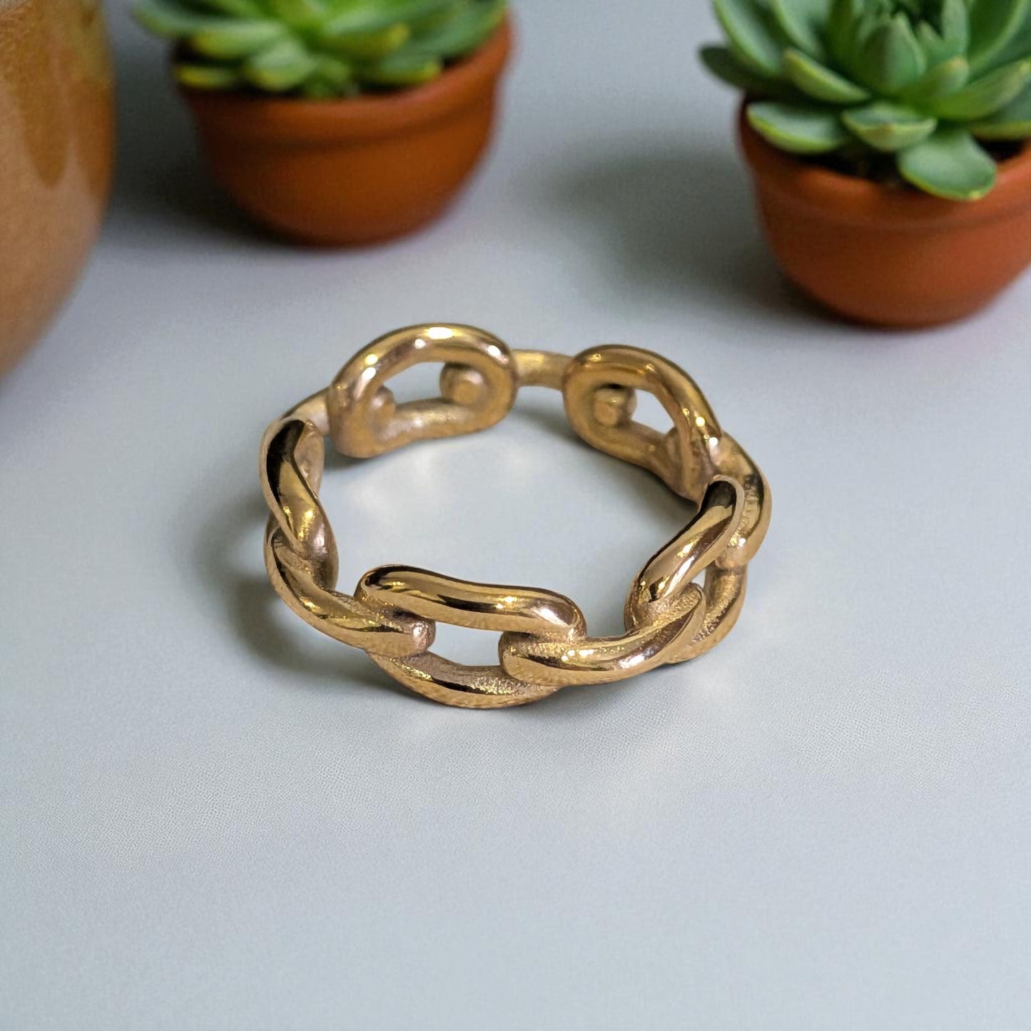 18k Gold plated chain-style ring on a light gray surface with potted plants in the background