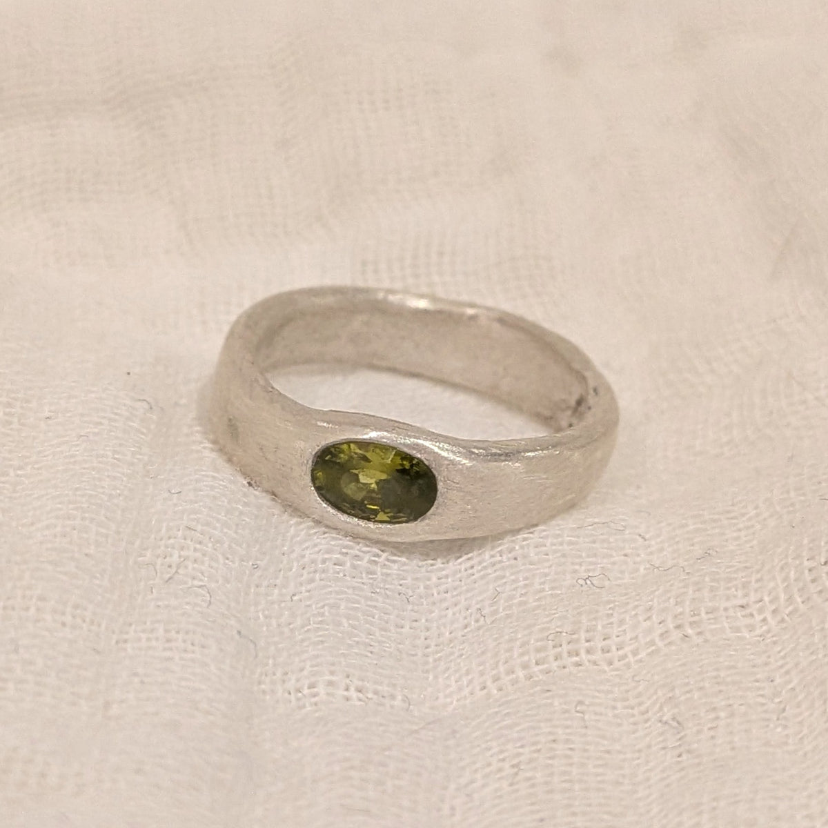 A rustic silver ring with a small oval green gemstone, resting on a textured off-white fabric background.