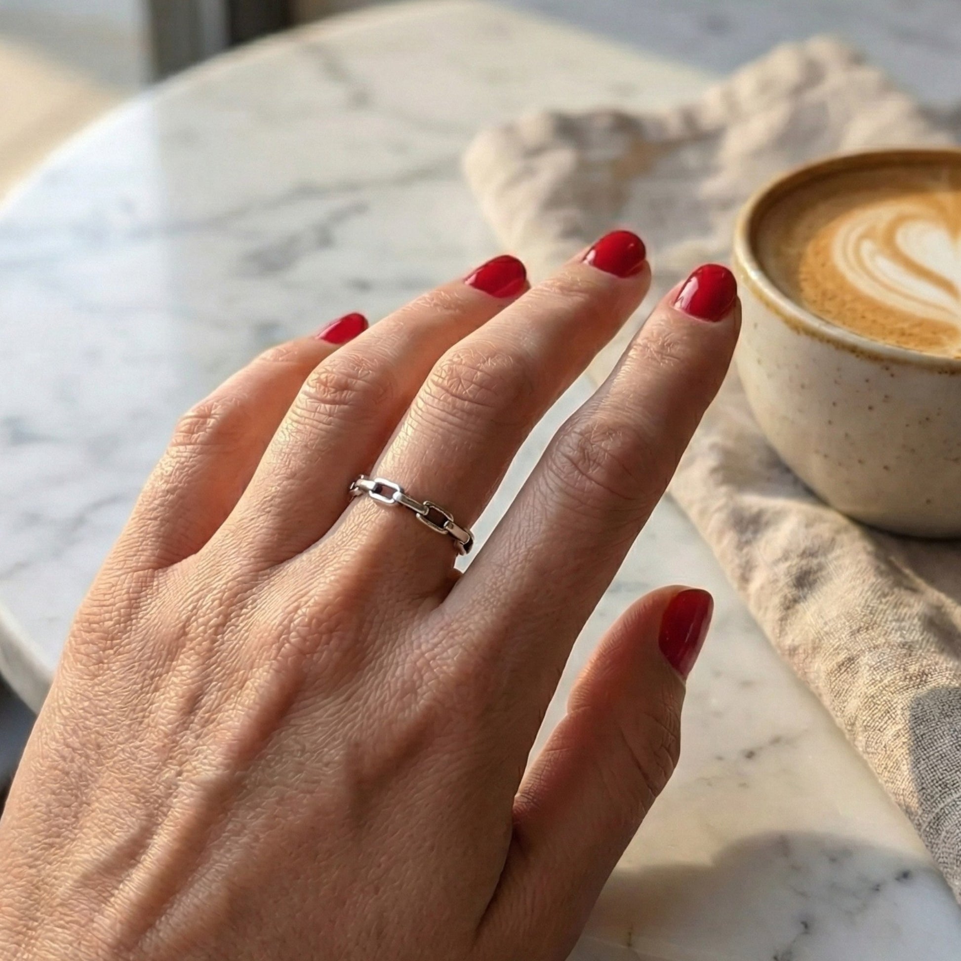 Hand with red nail polish wearing a silver ring on a marble surface with a coffee cup.